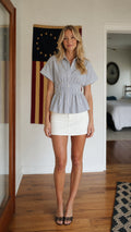 Front view of woman wearing blue striped button-down peplum top and white skirt
